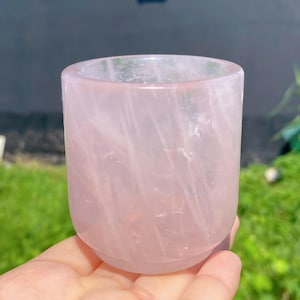 Puede incluir: Una taza de cuarzo rosa translúcido, sostenida con la mano. La taza es de color rosa suave con vetas naturales. Tiene una base redondeada y lados rectos, con un acabado liso y pulido. El fondo está borroso, con follaje verde y una superficie gris.