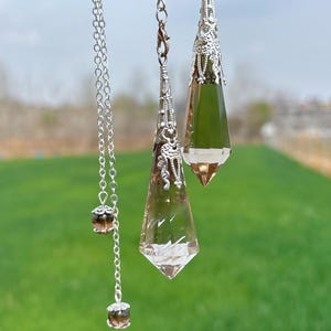 May include: Two clear quartz crystal pendulums with silver accents and chains. The larger pendulum is hanging from a silver chain with two brown beads. The smaller pendulum is hanging from a silver chain.