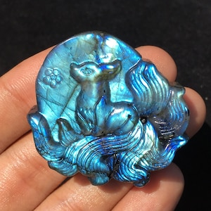 Op de afbeelding: Een blauwe labradorit-steenhouwwerk van een vos die op een maan zit met een bloemdessin. Het beeldhouwwerk is gedetailleerd en ingewikkeld, met een glinsterende, iriserende afwerking.