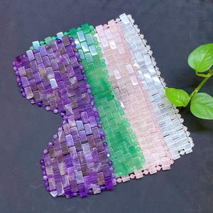 Puede incluir: Una máscara para los ojos de color morado, verde y blanco hecha de pequeñas cuentas de piedras preciosas rectangulares. La máscara está diseñada para colocarse sobre los ojos y está destinada a la relajación y la belleza.