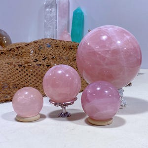 May include: Four pink rose quartz spheres of varying sizes on decorative stands. The spheres are smooth and polished, with a light pink hue.