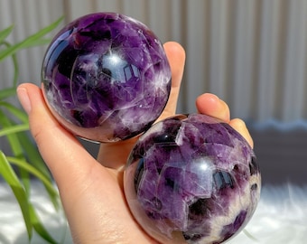 Op de afbeelding: Twee amethist kristallen bollen, één in de palm van een hand en de andere rustend op de hand. De bollen zijn diep paars van kleur met witte en zwarte insluitsels.