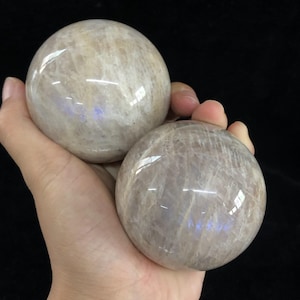 Puede incluir: Dos esferas de piedra lunar blanca iridiscente, cada una con una superficie lisa y pulida. Las esferas se sostienen en una mano, mostrando su tamaño y sus intrincados patrones.
