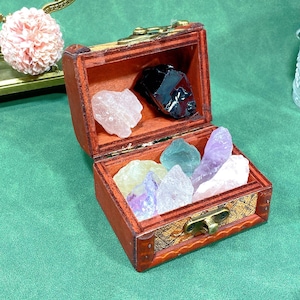 May include: A small, open wooden treasure chest filled with colourful crystals. The chest is reddish-brown with a gold-coloured clasp. Crystals include pink, black, yellow, green, and purple varieties. The chest sits on a green surface.
