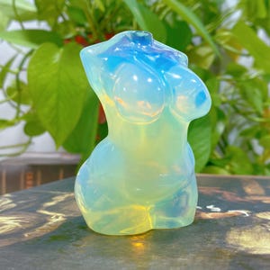 Puede incluir: Una escultura de torso femenino de cristal translúcido, iridiscente, azul claro y amarillo, tallada a mano y pulida.