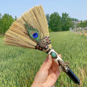 May include: A decorative broom with a natural straw brush head, decorated with a peacock feather and gemstone accents. The handle features a black crystal and ornate metalwork with a pentagram design. The broom is held against a backdrop of green fields.