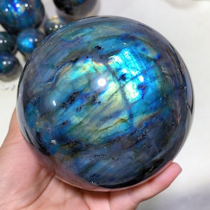 May include: A large, polished labradorite sphere with iridescent blue, green, and gold colours. The sphere is smooth and reflective, with a variety of patterns and textures.