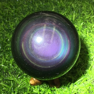 Puede incluir: Una esfera oscura iridiscente con un centro morado y un brillo arcoíris. La esfera está sentada sobre una superficie verde.