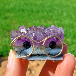 Può includere: Un'esclusiva formazione di cristalli di ametista con un paio di occhiali con montatura in filo d'oro. Gli occhiali hanno lenti nere. Il cristallo è di colore viola intenso con una base blu e bianca. Lo sfondo è verde sfocato.