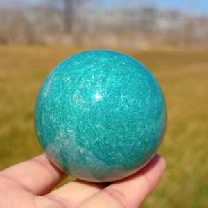 Puede incluir: Una esfera de amazonita verde pulida y lisa. La esfera tiene un patr&oacute;n de m&aacute;rmol verde claro y blanco.