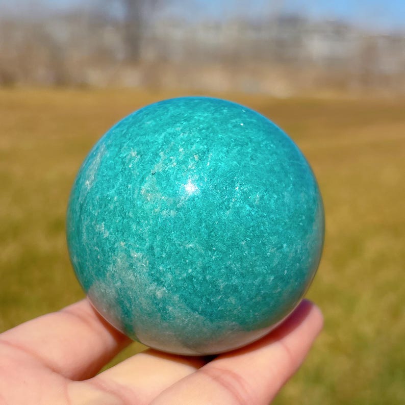Puede incluir: Una esfera de amazonita verde pulida y lisa. La esfera tiene un patr&oacute;n de m&aacute;rmol verde claro y blanco.