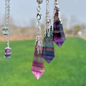 May include: Three silver and crystal pendulums with ornate detailing. The pendulums are made of fluorite and have a variety of colours, including purple, green, and pink.
