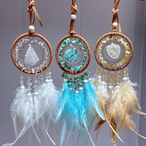 May include: Three dream catchers with brown hoops, decorated with crystals, beads, and feathers. The dream catchers have white, blue, and tan feathers, with crystals and stones in the centre of each hoop. Each is suspended by a leather cord.