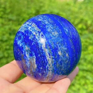 Peut inclure: Une sphère polie en lapis-lazuli, présentant une teinte bleu profond avec des stries blanches et des éclats dorés. La pierre précieuse est tenue dans une main, avec un arrière-plan vert flou.