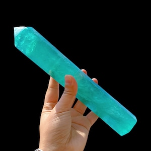 May include: A large, vibrant turquoise blue crystal point, also known as a wand, is held in a hand. The crystal is smooth and polished, with a pointed tip and a flat base.