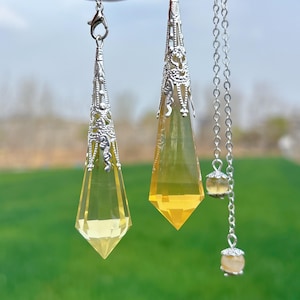 Pode incluir: Dois pêndulos de cristal amarelo com detalhes em filigrana prateada pendurados em correntes prateadas. Os pêndulos são pontiagudos na parte inferior e têm um design facetado.