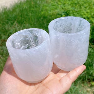 Puede incluir: Dos vasos de cristal de cuarzo transparente. Los vasos son cilíndricos con fondos redondeados. El cuarzo tiene un aspecto translúcido y lechoso con vetas internas visibles. Los vasos se sostienen en una mano, con un fondo verde.