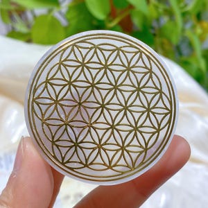 Könnte beinhalten: Weißer Selenitkristall mit einem goldenen Lebensblume-Design. Der Kristall ist rund und hat eine glatte Oberfläche.