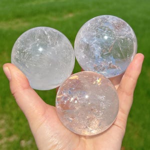 Pode incluir: Três esferas de cristal de quartzo transparente de tamanhos variados são seguradas em uma mão contra um fundo de grama verde. As esferas têm fraturas e inclusões internas, capturando a luz e criando um efeito brilhante. As esferas são populares na comunidade espiritual.