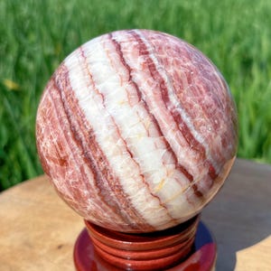 Op de afbeelding: Een gepolijste bolvormige steen met afwisselende banden van diep bordeauxrode, roodbruine en doorschijnend witte minerale lagen. De bol staat op een ronde, donkerroodbruine houten standaard en heeft een glad, reflecterend oppervlak.