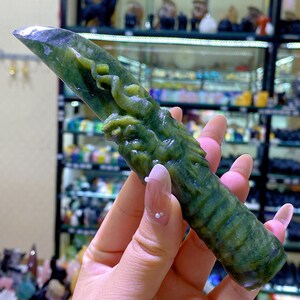 May include: A green jade carving of a dragon. The jade is a mottled green color with darker green details. The dragon is carved in relief, with its head and body visible. The carving is held in a person's hand.