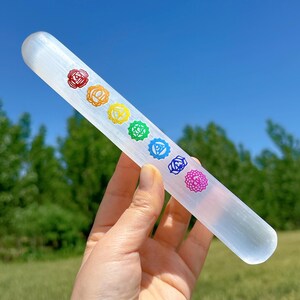 Puede incluir: Una varita de selenita transparente, de unos 15 cm de largo, con siete símbolos de chakra de colores. La varita se sostiene sobre un fondo de cielo azul y árboles verdes.