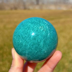 Puede incluir: Una esfera de amazonita verde lisa y pulida. La esfera tiene un color verde claro con vetas verde oscuro en toda su superficie. La esfera tiene aproximadamente el tama&ntilde;o de una pelota de b&eacute;isbol.