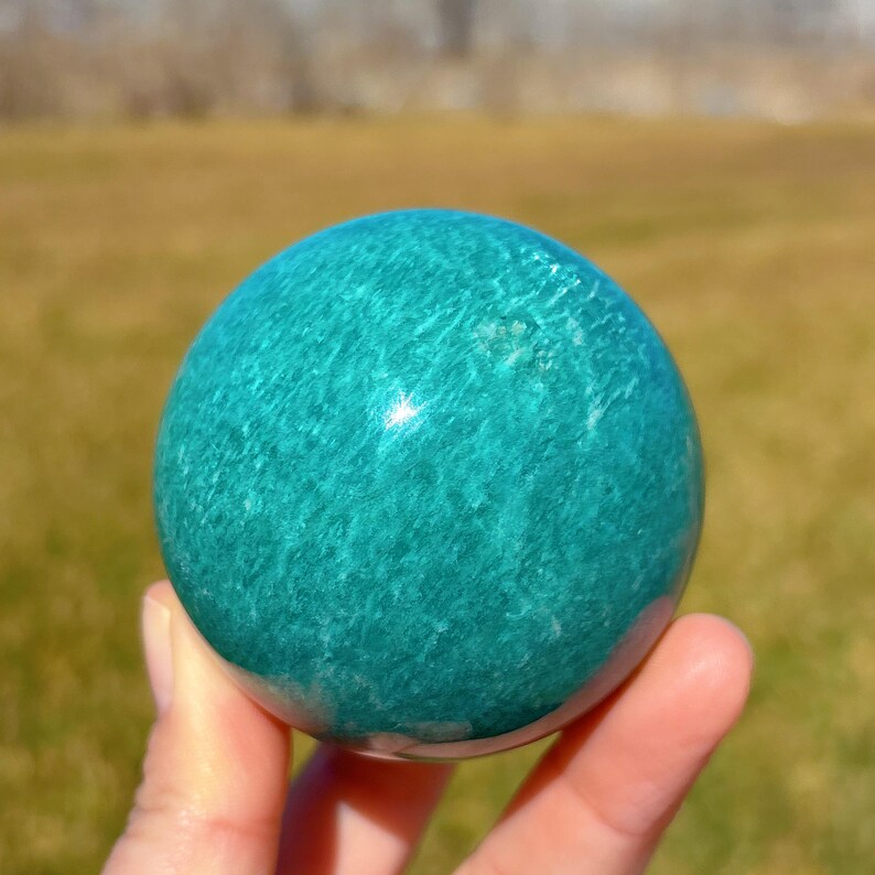 Puede incluir: Una esfera de amazonita verde lisa y pulida. La esfera tiene un color verde claro con vetas verde oscuro en toda su superficie. La esfera tiene aproximadamente el tama&ntilde;o de una pelota de b&eacute;isbol.