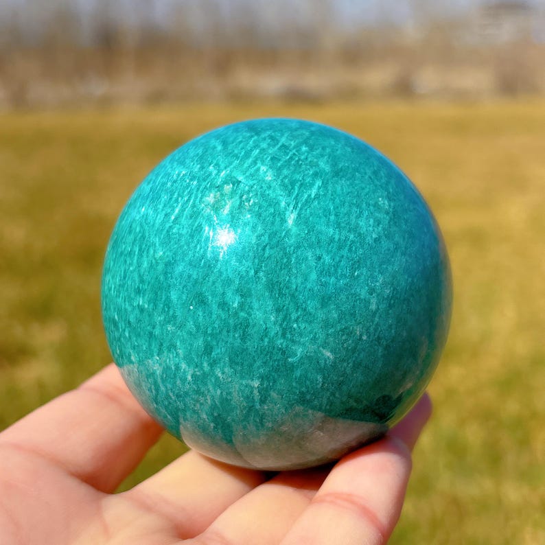 Puede incluir: Una esfera de amazonita verde lisa y pulida. La esfera es de un verde vibrante con vetas blancas en todo.