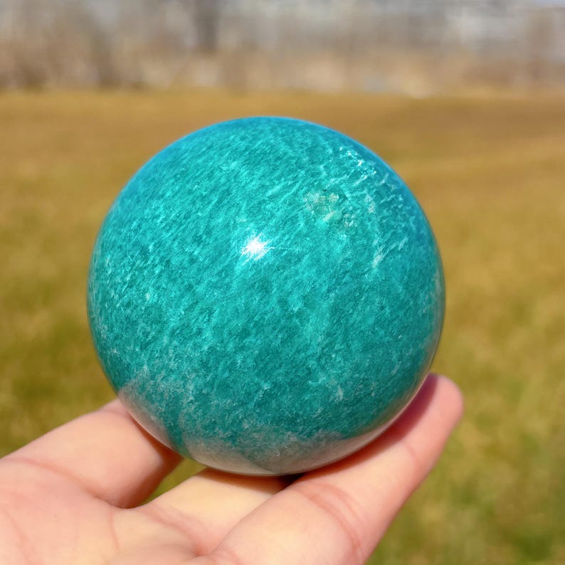 Puede incluir: Una esfera de amazonita lisa y verde, pulida a alto brillo. La esfera es aproximadamente del tama&ntilde;o de una pelota de golf y tiene un patr&oacute;n de remolino de verde claro y oscuro.