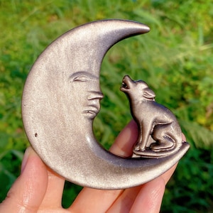 Può includere: Scultura metallica color bronzo raffigurante una luna crescente con un volto scolpito e un lupo che ulula. Lo sfondo sfocato è verde, suggerendo vegetazione.