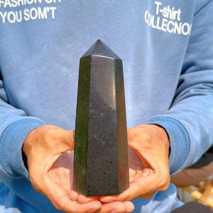 May include: A black obsidian crystal point, also known as a black obsidian tower, is a dark, glassy, and pointed crystal. The crystal is being held in a person's hands.