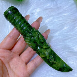 May include: A hand-carved green jade knife with a dragon design. The knife features a detailed dragon carving on the handle and blade. The jade has varying shades of green, creating a unique and natural appearance. The knife is held against a white, fluffy background.