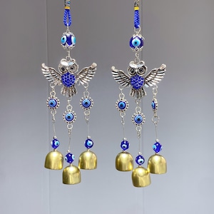 May include: Two hanging decorative charms featuring silver owl figures with blue accents and wings. Each charm has multiple strands of blue evil eye beads and small gold bells. The charms are suspended from blue braided cords.