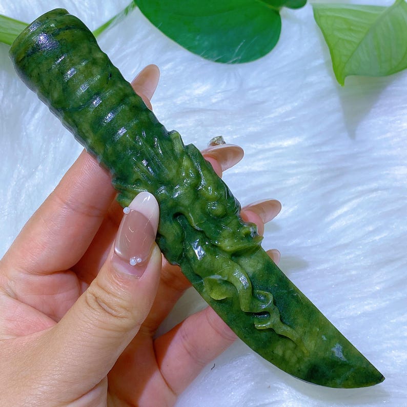 May include: A carved jade dagger with a dragon motif. The dagger is a deep green color and features intricate carvings along the handle and blade. The dragon is sculpted in relief, adding texture and detail to the piece. The blade is smooth and polished.