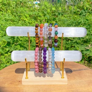 Op de afbeelding: Een displaystandaard met twee witte horizontale staven houdt verschillende armbanden met kralen vast. De armbanden hebben een verscheidenheid aan kleuren en edelstenen, waaronder tijgeroog, rozenkwarts en amethist. De standaard heeft goudkleurige steunen en een houten basis.