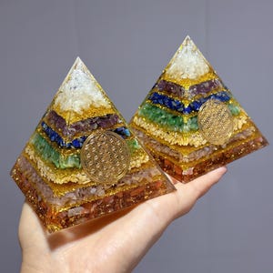 May include: Two orgone pyramids with layered colorful stones and gold glitter. Each pyramid features a gold-colored Flower of Life symbol. The pyramids are held in a hand, showcasing the intricate details and vibrant colors.