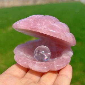 May include: A pink rose quartz clam shell with a clear crystal sphere inside. The clam shell is carved with detailed ridges and has a smooth, polished finish. The crystal sphere reflects light, creating a rainbow effect.