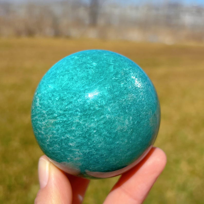 Puede incluir: Una esfera de amazonita verde pulida y lisa. La esfera tiene un color verde claro con vetas blancas.