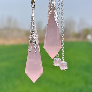 Könnte beinhalten: Zwei rosa Quarzkristall-Pendel mit silbernen Filigran-Akzenten und Ketten. Die Pendel hängen vor einem grünen Gras Hintergrund.