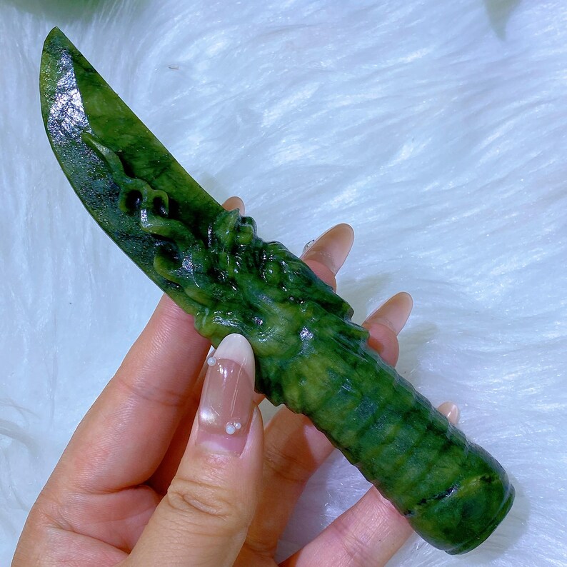 May include: A green jade knife with a detailed dragon carving on the handle. The blade is smooth and polished, while the handle features intricate textures. The jade has varying shades of green, creating a natural, organic look.