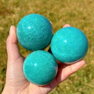 Puede incluir: Tres esferas lisas de amazonita verde, cada una del tama&ntilde;o de una pelota de golf, se sostienen en una mano. Las esferas est&aacute;n pulidas y tienen un brillo sutil.