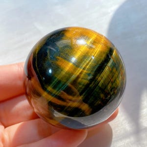 May include: A polished tiger's eye sphere with a mix of golden yellow, brown, and blue-green hues. The sphere is smooth and reflective, showcasing the natural patterns of the stone.
