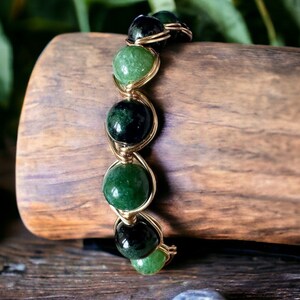 Puede incluir: Un brazalete de piedras preciosas verdes y negras con alambre dorado envuelto. El brazalete está hecho con cuentas redondas y está sobre una superficie de madera.