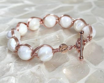 Puede incluir: Pulsera de oro rosa con cuentas blancas redondas. Las cuentas están envueltas con alambre de oro rosa. La pulsera tiene un cierre de palanca y la palabra "Essel" está grabada en el cierre.