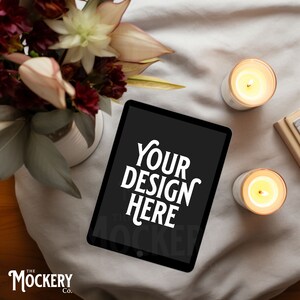Puede incluir: Una tableta negra con una pantalla blanca que muestra el texto "YOUR DESIGN HERE". La tableta está sobre una superficie de tela blanca con dos velas encendidas y un ramo de flores.