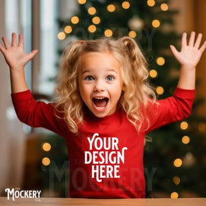 Op de afbeelding: Een jong meisje draagt een rood shirt met lange mouwen met de tekst "Your Design Here" op de voorkant. Ze glimlacht en heeft haar armen in de lucht. De achtergrond is een wazig beeld van een kerstboom met lichtjes.