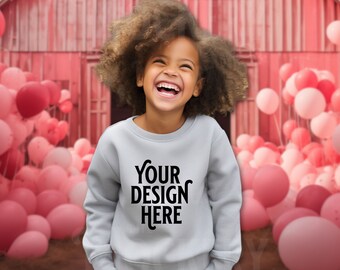 Może przedstawiać: Młoda osoba w szarej bluzie z nadrukiem "YOUR DESIGN HERE" na przodzie. Osoba uśmiecha się i stoi przed czerwonym stodołą z różowymi balonami.