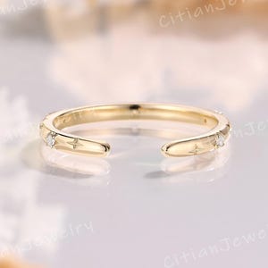 May include: A gold open ring featuring a polished band with small engraved star motifs and tiny inset clear stones. The ring has a gap at the front, allowing for an adjustable fit, and displays a smooth, reflective metallic finish throughout.