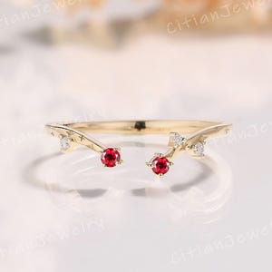 May include: A gold-colored ring with a unique open design. The ring features two bright red gemstones and several small, clear gemstones. The ring has a delicate, elegant appearance, perfect for special occasions. The ring is a beautiful piece of jewelry.